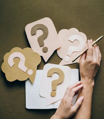 Hands holding pen next to paper question marks on notebook, symbolizing inquiry and creativity.